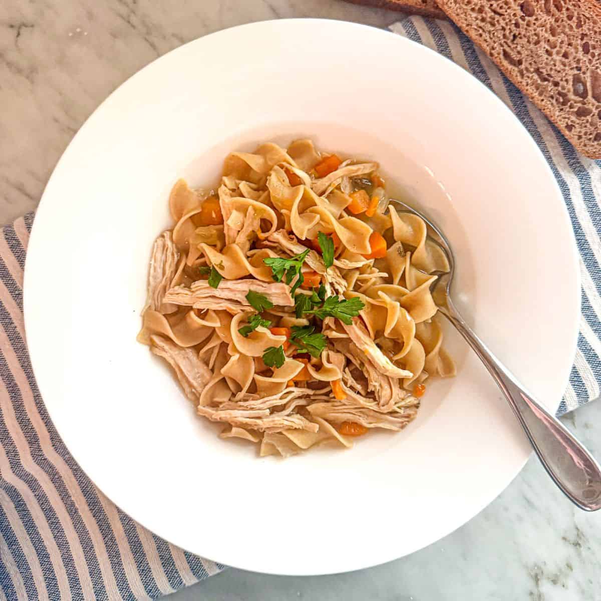 Savory Mediterranean diet chicken noodle soup with vegetables and fresh herbs in a white bowl on a marble surface.