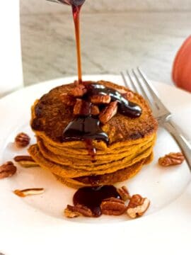A stack of Mediterranean Diet Kodiak Pumpkin Pancakes on a white plated. Topped with nuts and with maple syrup being poured over them.