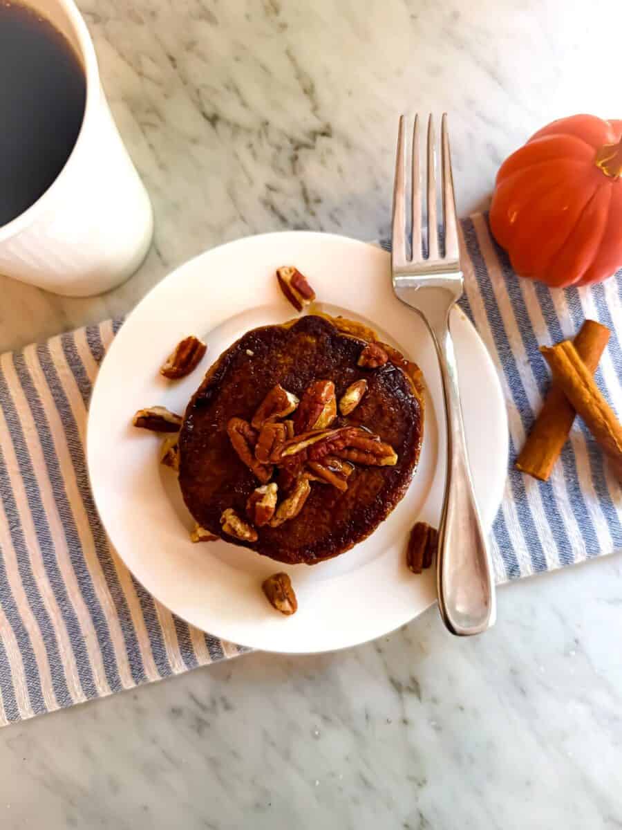 Mediterranean diet Kodiak pumpkin pancakes topped with pecan nuts and maple syrup. They are on a white plate with a white fork with a blue and white napkin, cup of coffee, decorative pumpkin and cinnamon stick nearby.