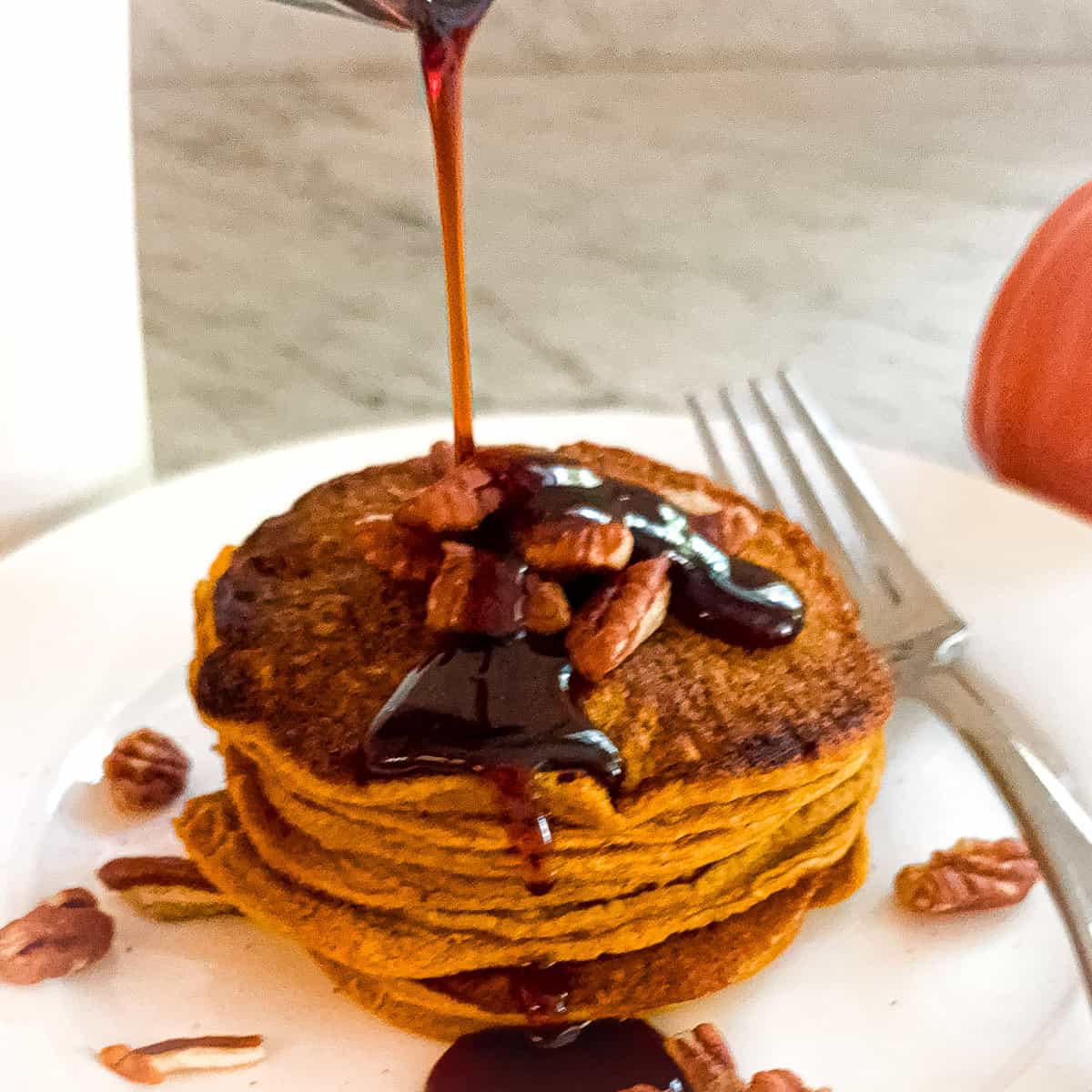 A stack of Mediterranean Diet Kodiak Pumpkin Pancakes on a white plated. Topped with nuts and with maple syrup being poured over them.