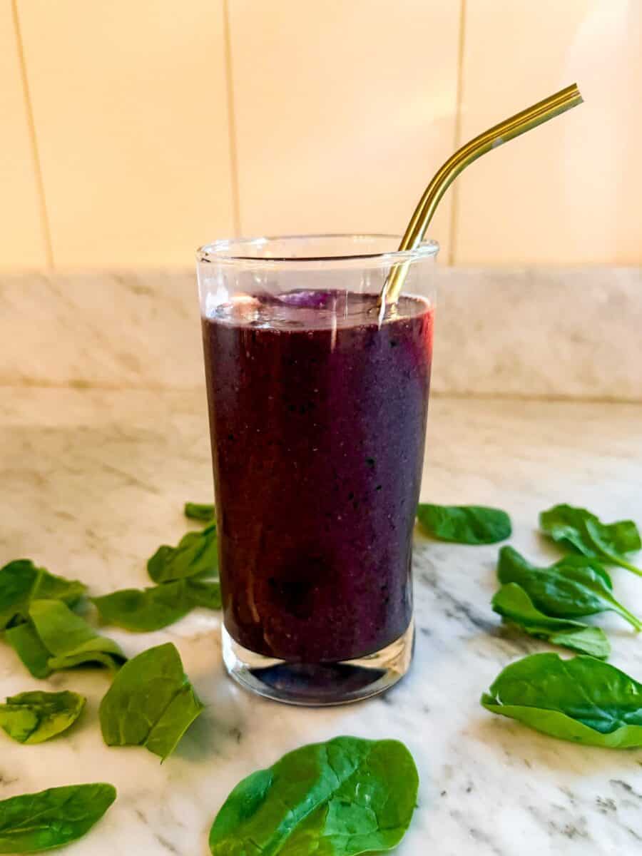 A Mediterranean Diet smoothie containing spinach, blueberries, and flax in a glass on a marble countertop. Surrounded by spinach leaves and with a metal straw.