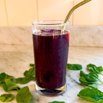 A Mediterranean Diet smoothie containing spinach, blueberries, and flax in a glass on a marble countertop. Surrounded by spinach leaves and with a metal straw.