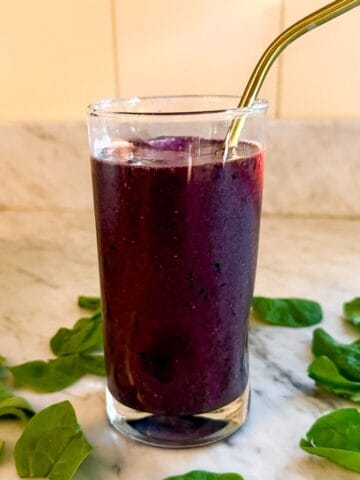 A Mediterranean Diet smoothie containing spinach, blueberries, and flax in a glass on a marble countertop. Surrounded by spinach leaves and with a metal straw.