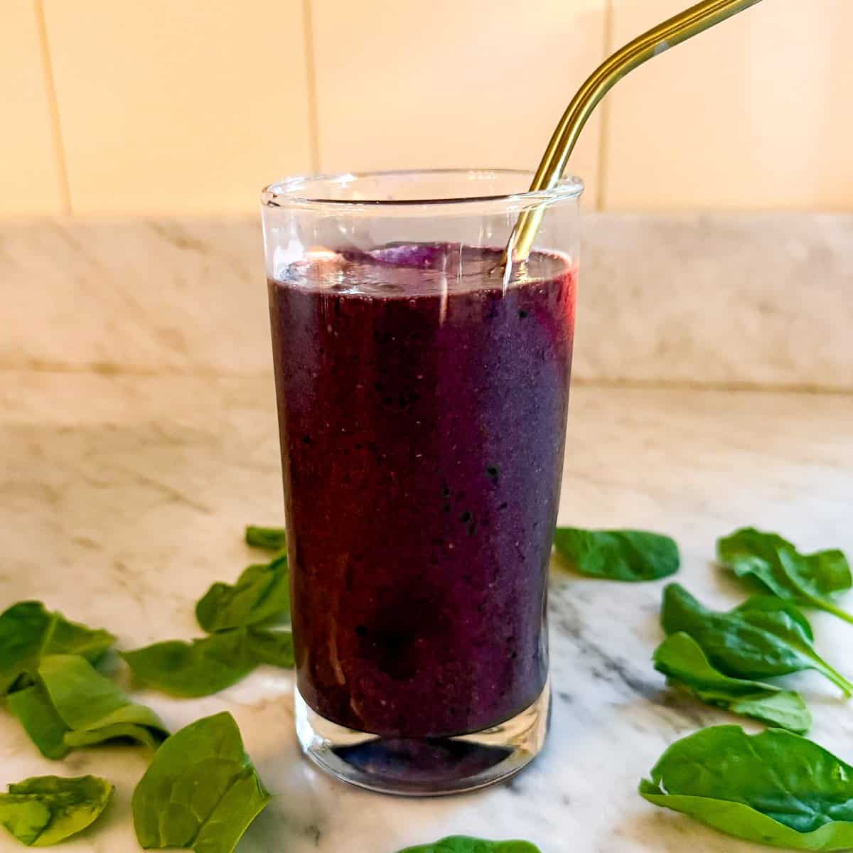 A Mediterranean Diet smoothie containing spinach, blueberries, and flax in a glass on a marble countertop. Surrounded by spinach leaves and with a metal straw.