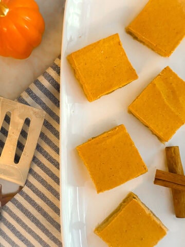 Pumpkin pie bars on a white plate resting on a blue and white dishtowel on a marble countertop. Flanked by a ceramic pumpkin, cinnamon sticks, and a silver spatula