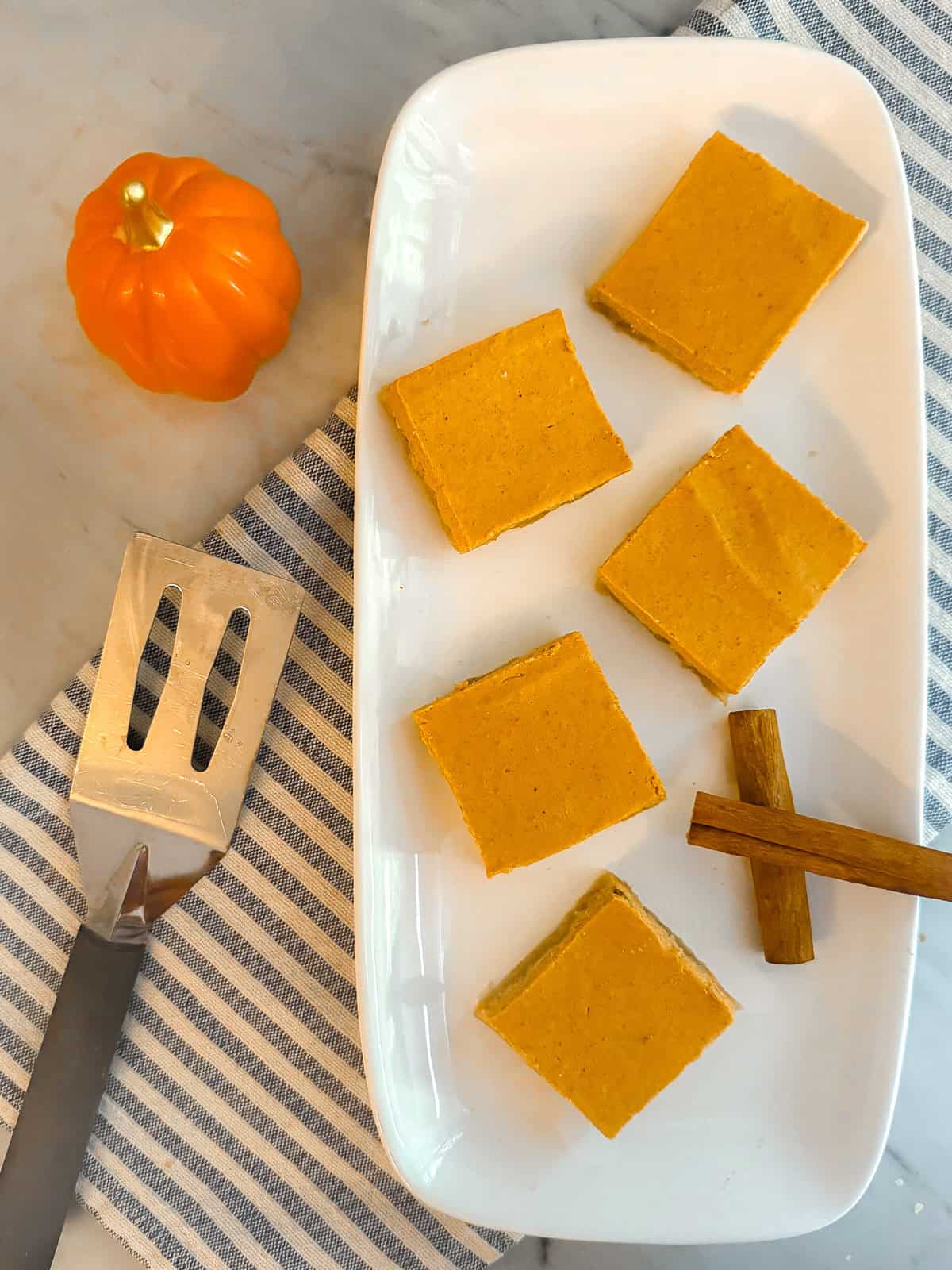 Pumpkin pie bars on a white plate resting on a blue and white dishtowel on a marble countertop. Flanked by a ceramic pumpkin, cinnamon sticks, and a silver spatula