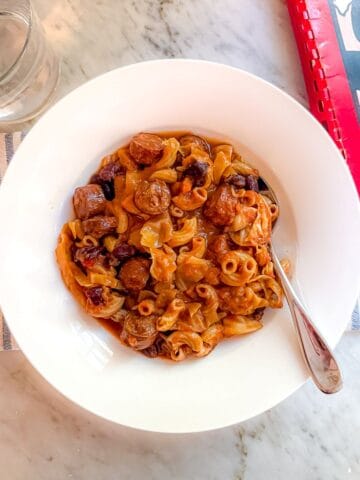 A healthier version of Portuguese Bean Soup that is Mediterranean Diet friendly in a white bowl with a spoon atop a marble countertop. A cookbook and a glass of water are nearby.