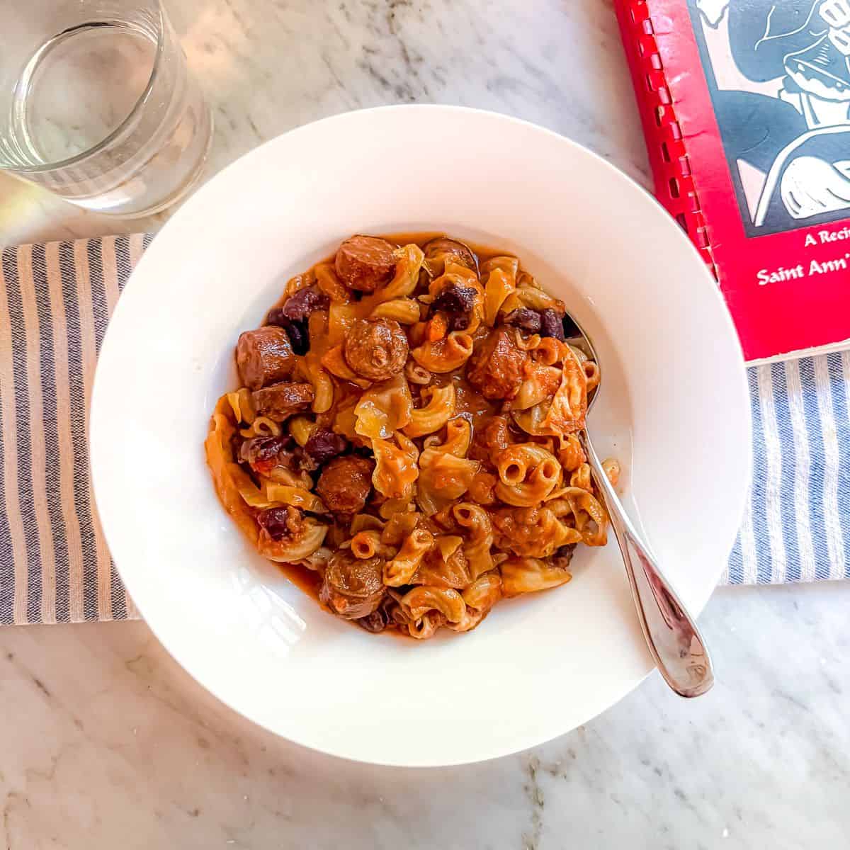 A healthier version of Portuguese Bean Soup that is Mediterranean Diet friendly in a white bowl with a spoon atop a marble countertop. A cookbook and a glass of water are nearby.