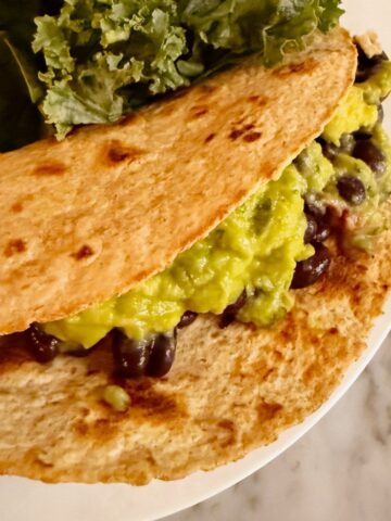 A vegan taco with black beans, guacamole, and a whole wheat tortilla. Placed on a plate with kale and mango.