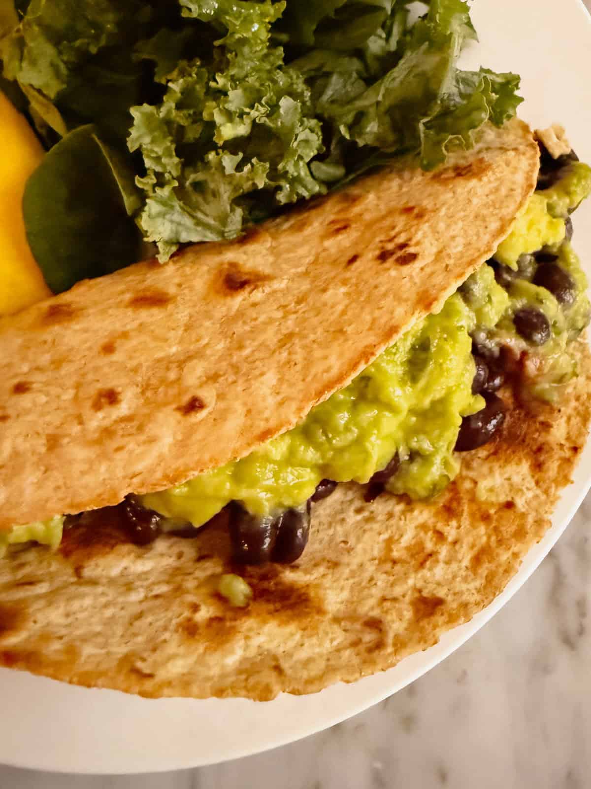 A vegan taco with black beans, guacamole, and a whole wheat tortilla. Placed on a plate with kale and mango.