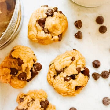 Dairy Free Chocolate Chip Cookies on a marble countertop surrounded by chocolate chips and a cup of milk