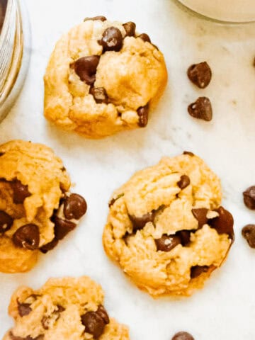Dairy Free Chocolate Chip Cookies on a marble countertop surrounded by chocolate chips and a cup of milk