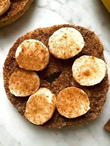 A Mediterranean Diet breakfast consisting of a whole wheat bagel with almond butter topped with ground flaxseed, banana slices and a sprinkle of cinnamon on a marble surface. Nearby are cinnamon sticks and a part of a banana.