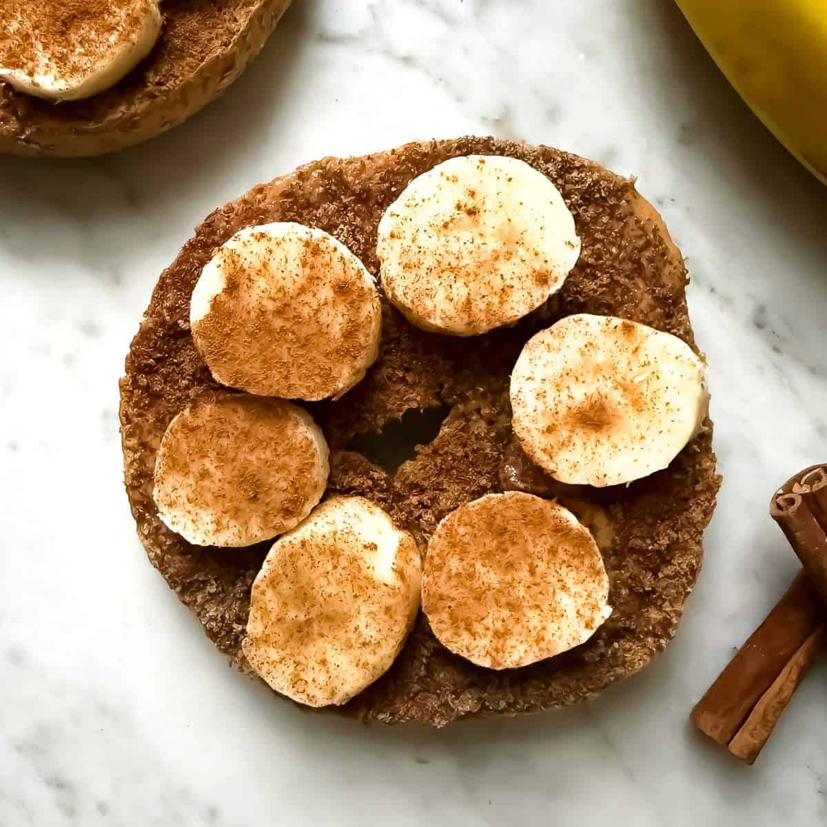 A Mediterranean Diet breakfast consisting of a whole wheat bagel with almond butter topped with ground flaxseed, banana slices and a sprinkle of cinnamon on a marble surface. Nearby are cinnamon sticks and a part of a banana.