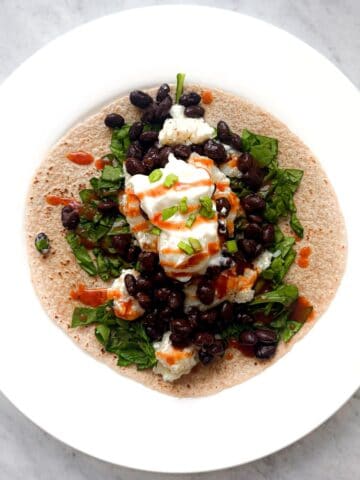 A whole wheat tortilla topped with black beans, fresh spinach, egg whites, a dollop of sour cream, and taco sauce, garnished with green onions.