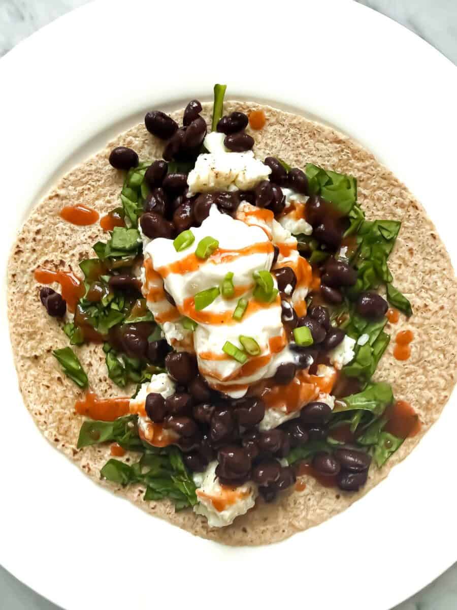 Healthy Breakfast taco made of whole wheat tortilla, egg whites, black beans, spinach, green onion, Greek yogurt on a white plate.