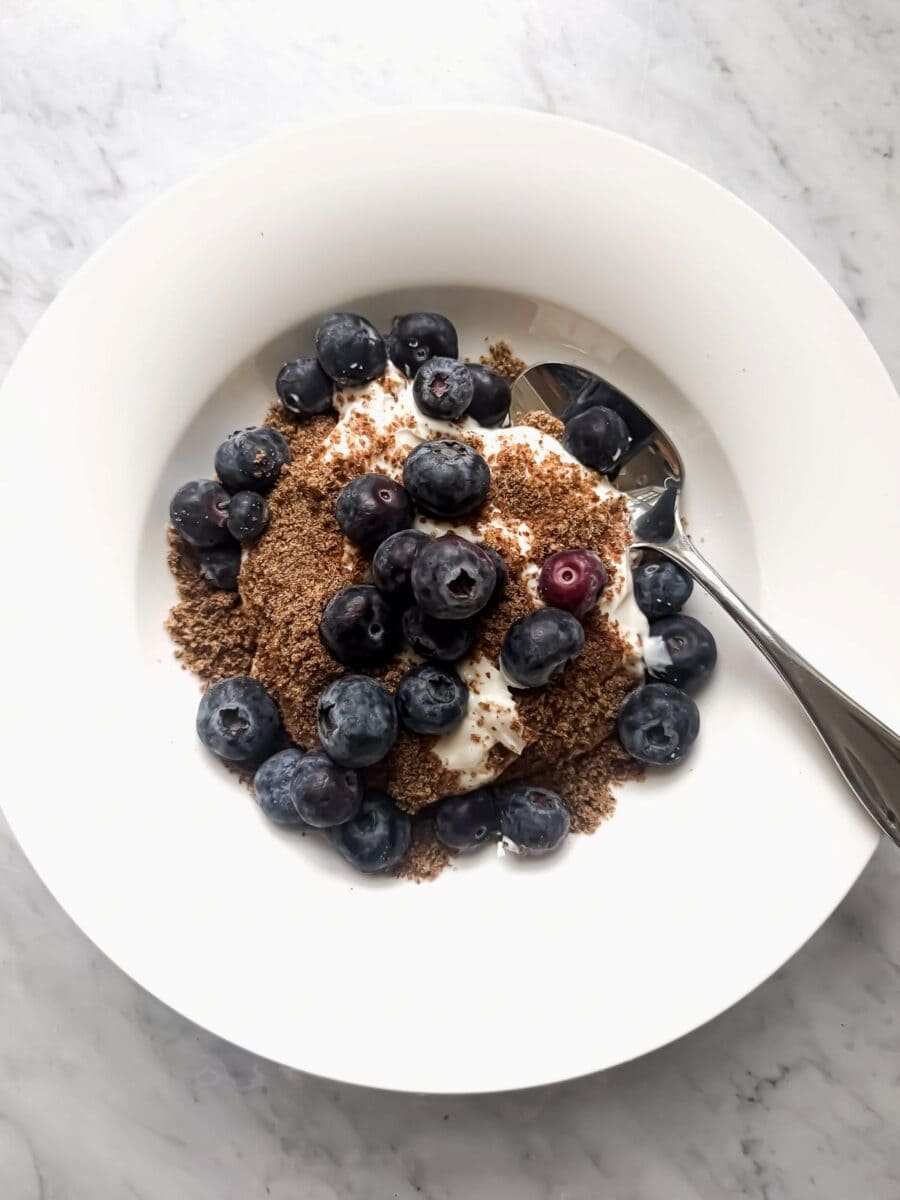 A high protein yogurt bowl with Greek yogurt, ground flaxseed, and blueberries in a white bowl on a marble counter. A spoon rests in the bowl