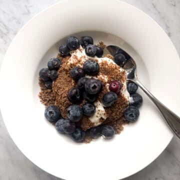 A white bowl filled with a Mediterranean Diet Breakfast. Creamy nonfat Greek yogurt, topped with blueberries and a sprinkle of ground flaxseed, with a spoon on the side. Clean, fresh, and inviting.