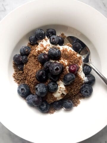 A white bowl filled with a Mediterranean Diet Breakfast. Creamy nonfat Greek yogurt, topped with blueberries and a sprinkle of ground flaxseed, with a spoon on the side. Clean, fresh, and inviting.