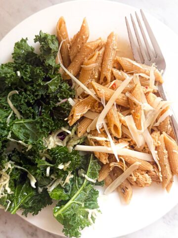 Crock Pot Garlic Parmesan Chicken Pasta on a plate with a simple salad. A fork is resting on the plate. The plate is on a marble countertop.