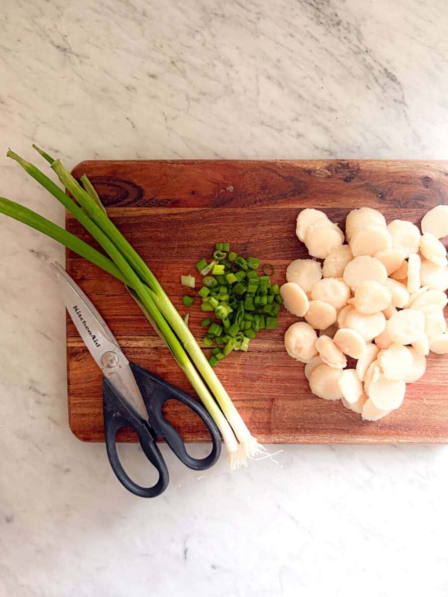 Scissors, green onions, and water chestnuts on a cutting board atop a marble countertop