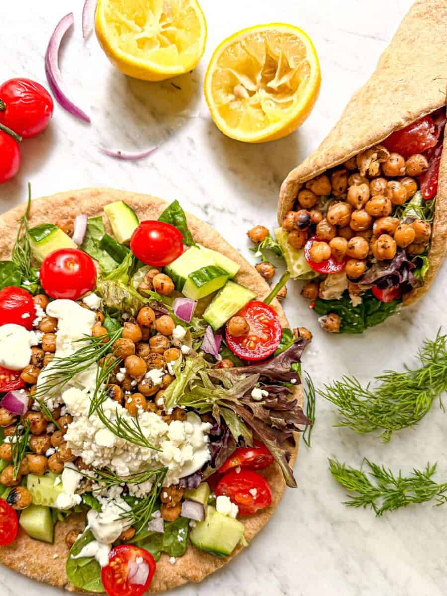 Open whole grain pita topped with marinated chickpeas, mixed greens, tomatoes, cucumber, red onion, feta, tzatziki, and fresh dill beside an assembled wrap.