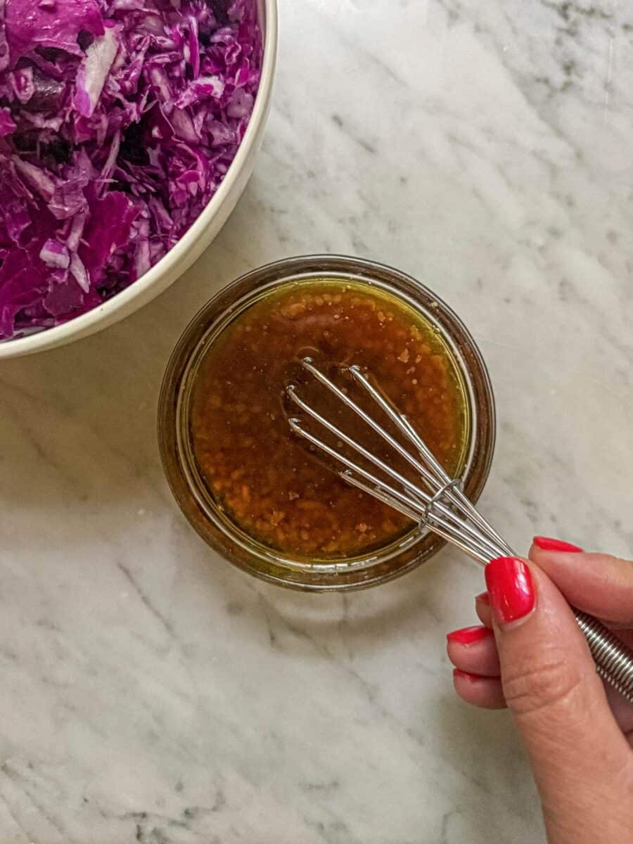 Sesame ginger dressing being whisked in a small jar beside shredded red cabbage.
