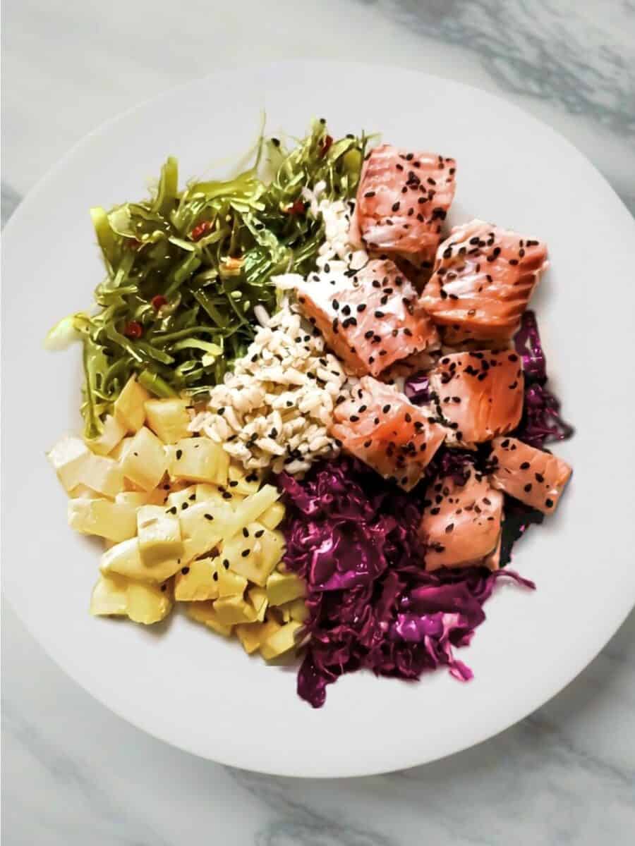 Cooked salmon poke bowl with brown rice, mango, seaweed salad, sesame ginger red cabbage, and black sesame seeds
