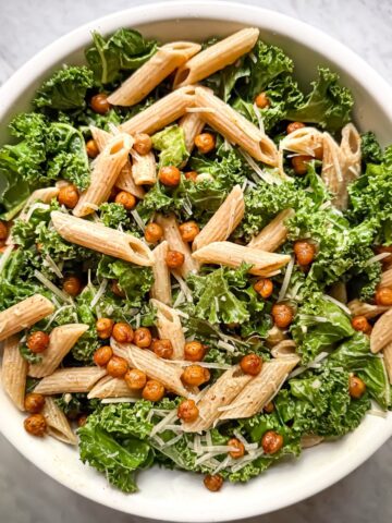Kale pasta salad with crispy chickpeas in a white bowl. The salad contains small pieces of kale, crispy chickpeas seasoned with paprika, parmesan, whole wheat penne, and a bright lemon evoo dressing.