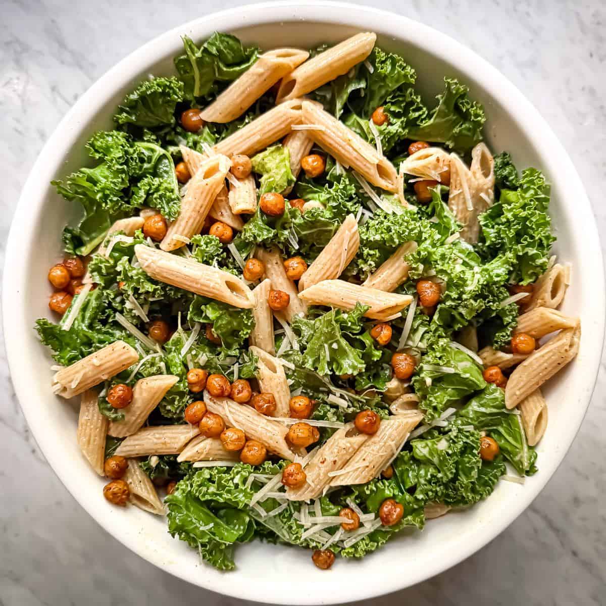 Kale pasta salad with crispy chickpeas in a white bowl. The salad contains small pieces of kale, crispy chickpeas seasoned with paprika, parmesan, whole wheat penne, and a bright lemon evoo dressing.