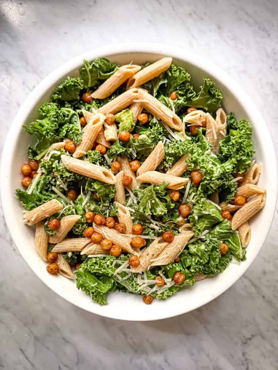 Kale pasta salad with crispy chickpeas in a white bowl. The salad contains small pieces of kale, crispy chickpeas seasoned with paprika, parmesan, whole wheat penne, and a bright lemon evoo dressing.