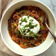 White bowl of vegan sweet potato black bean chili with vegan sour cream, cilantro, and green onion, with a spoon and a beige napkin on a marble surface