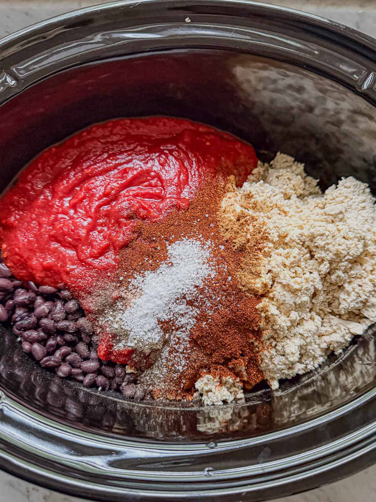 Slow cooker filled with crushed tomatoes, black beans, crumbled tofu, and chili spices, ready to cook vegan sweet potato chili