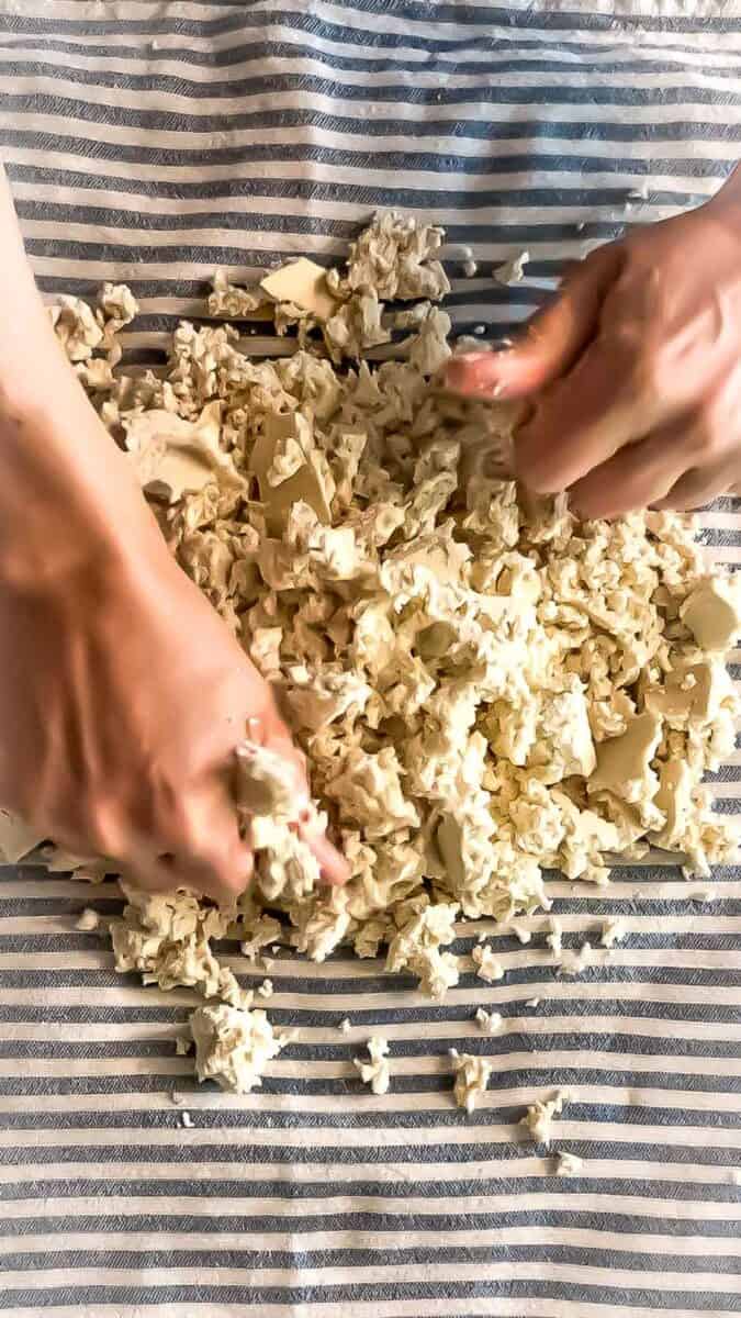 Hands crumbling a block of tofu into small pieces on a striped kitchen towel
