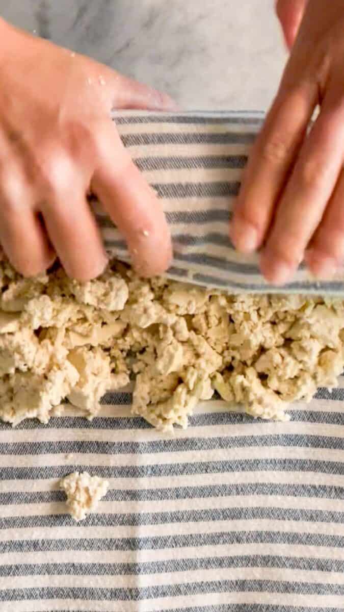 Crumbled tofu being pressed with a striped towel to squeeze out extra moisture