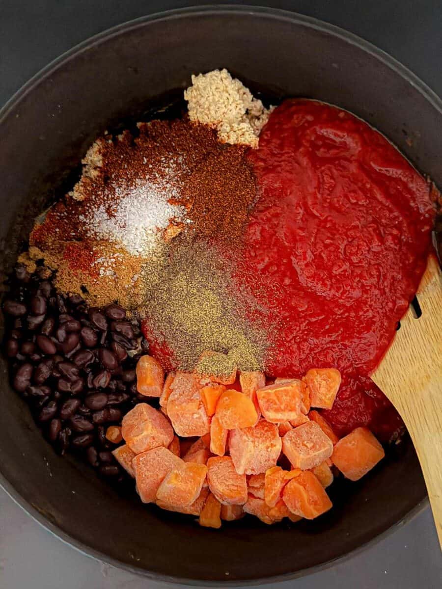 Dutch oven with black beans, frozen sweet potato cubes, crushed tomatoes, tofu crumbles, and spices added before stirring