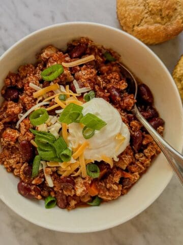 Close view of vegan tofu and kidney bean chili topped with shredded cheese, a creamy white topping, and green onions, with a spoon in the bowl.
