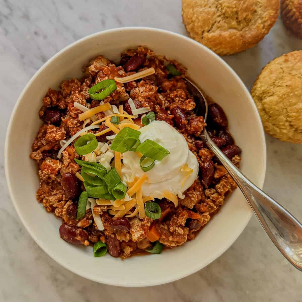 Close view of vegan tofu and kidney bean chili topped with shredded cheese, a creamy white topping, and green onions, with a spoon in the bowl.