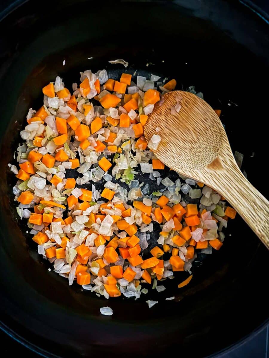 Diced onion, celery, and carrots in a black pot with a wooden spoon.