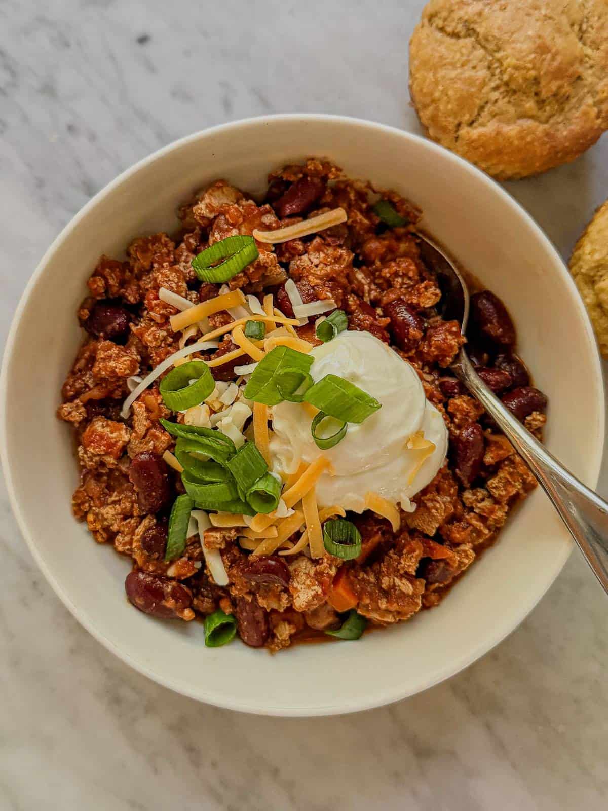 Close view of vegan tofu and kidney bean chili topped with shredded cheese, a creamy white topping, and green onions, with a spoon in the bowl.