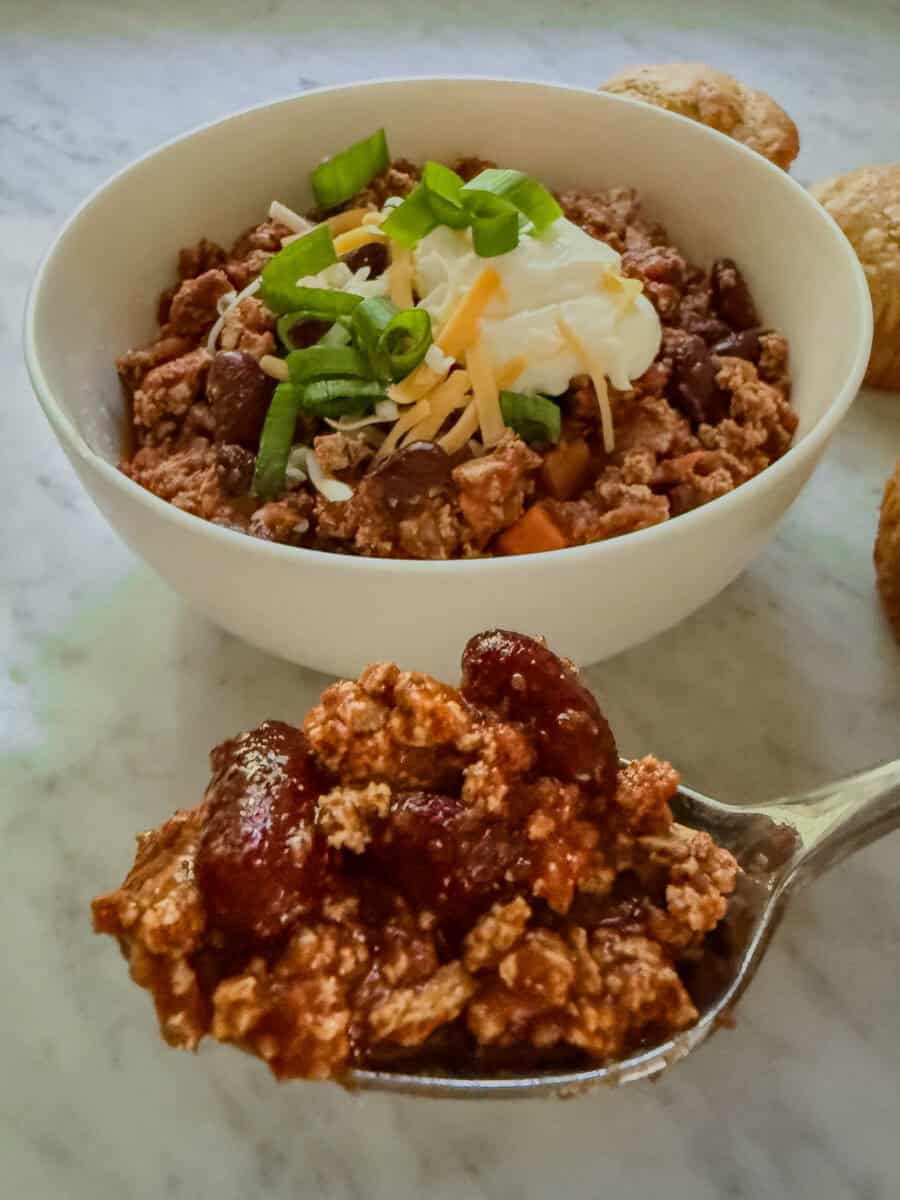 Spoonful of vegan chili with crumbled tofu and kidney beans in front of a bowl topped with shredded cheese, green onions, and a creamy white topping.